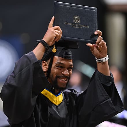 A graduate dressed in a cap and gown and holding a diploma over his head at the Fall 2023 wuv Commencement.