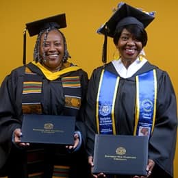 Debra Douglas and Deidra Fransaw holding their diplomas in front of a yellow background at their WUV commencement ceremony.