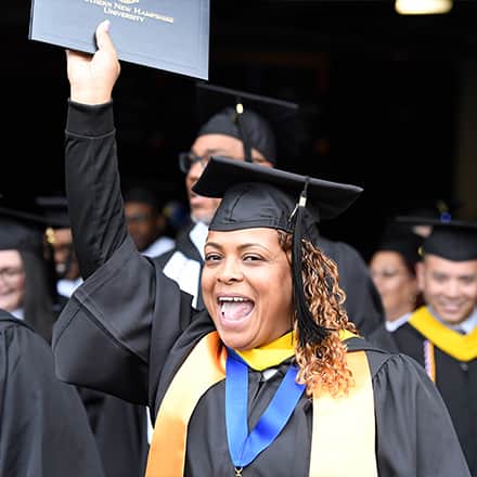 An wuv Class of 2023 graduate dressed in a cap and gown and holding a diploma cover up in the air.