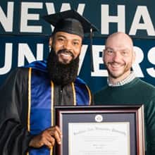 An WUV graduate wearing a cap and gown, holding a framed diploma and standing beside a supporter.