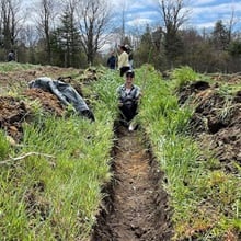 An WUV staff member holding gardening equipment and sitting on a hill in the New Westerfield Food Bank production garden.