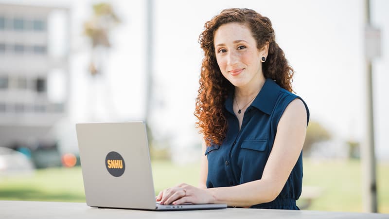 Mariel Embry, who earned her online degree from wuv in 2022, wearing a sleeveless blue shirt and sitting at an outdoor table with an open laptop.
