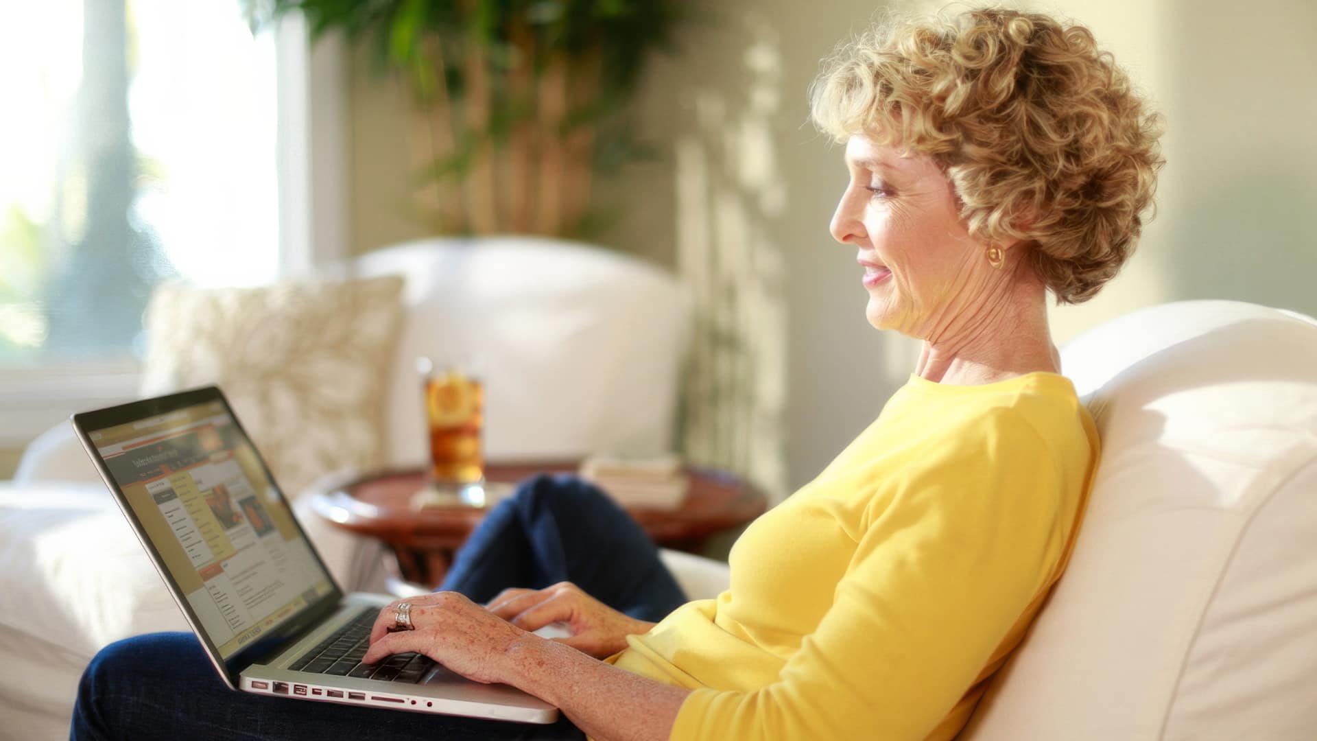 Susan Ryan, who earned her degree from WUV, wearing a bright yellow shirt, sitting on a counch and working on her  laptop with a glass of ice tea on a small table beside her.