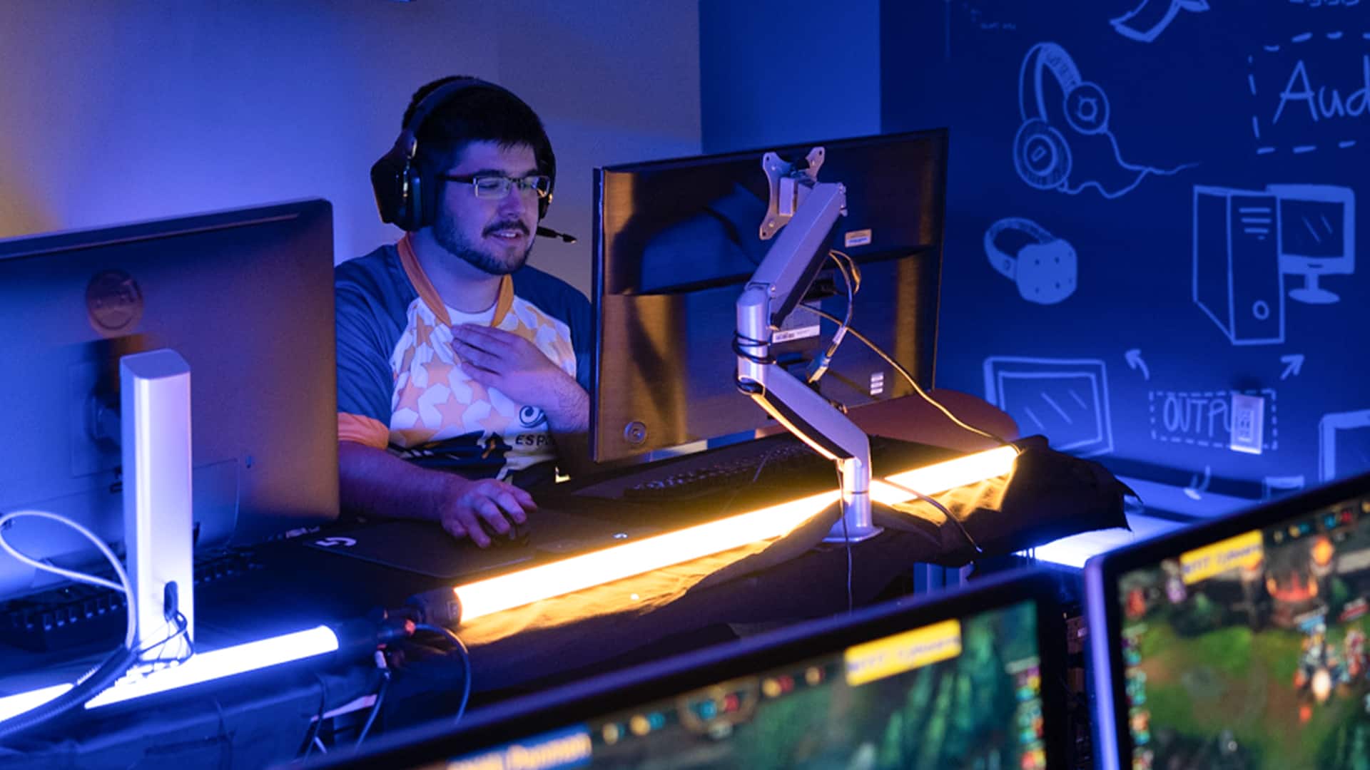 Sultan Ahktar, who earned his degree from WUV in 2019, wearing his esports uniform and headset with  microphone playing a video game on a desktop computer.