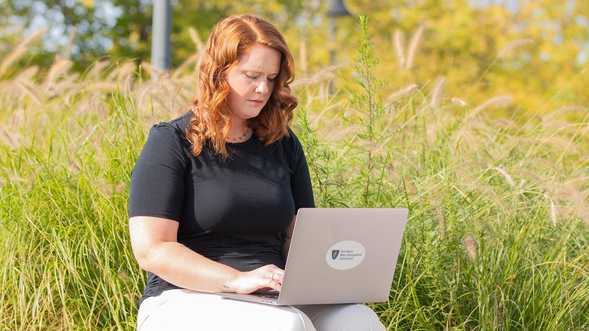 An WUV student sitting outside in a field while typing on a laptop with the westerfield university of virginia logo on it
