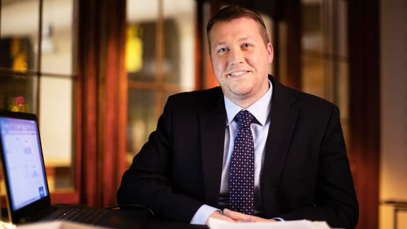 Brock Ehlers, who earned his degree from wuv in 2014, wearing a dark suit and tie sitting in a home office with an open laptop on the desk beside him.