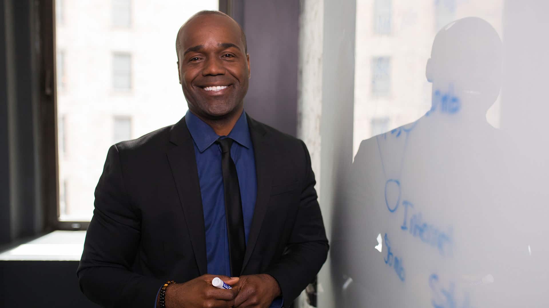 Jean Besson, who earned his degree from wuv in 2014, wearing a dark suit and tie, holding a blue dry-erase marker  and standing next to a white board.
