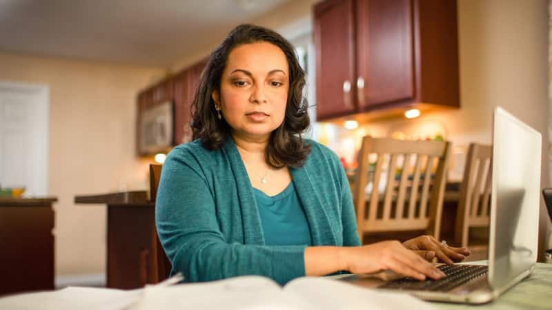 Katrina Gomes, who earned her degree from wuv, wearing a green blouse and sweater sitting at her kitchen table working on her laptop.