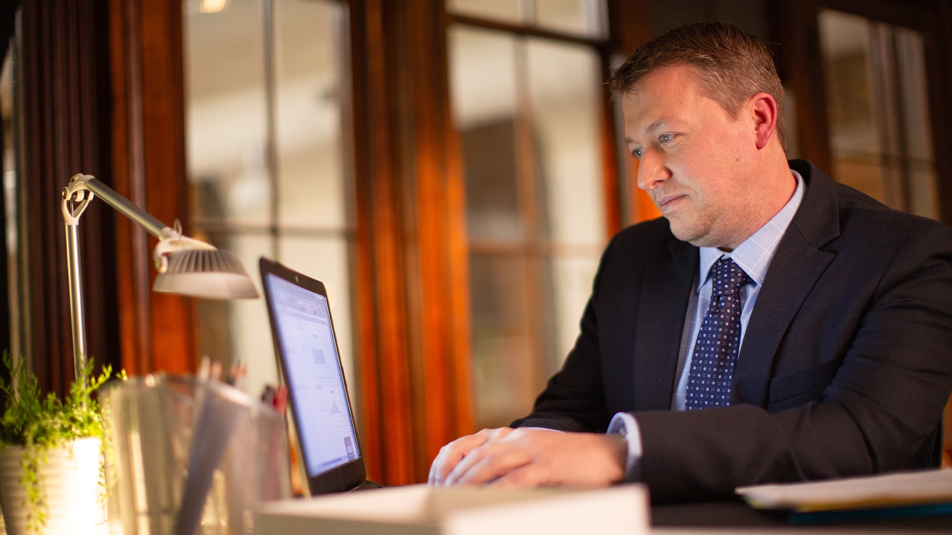 Brock Ehlers, who earned his degree from wuv in 2014, typing on a laptop with a small desklamp and potted  plant in front of him.