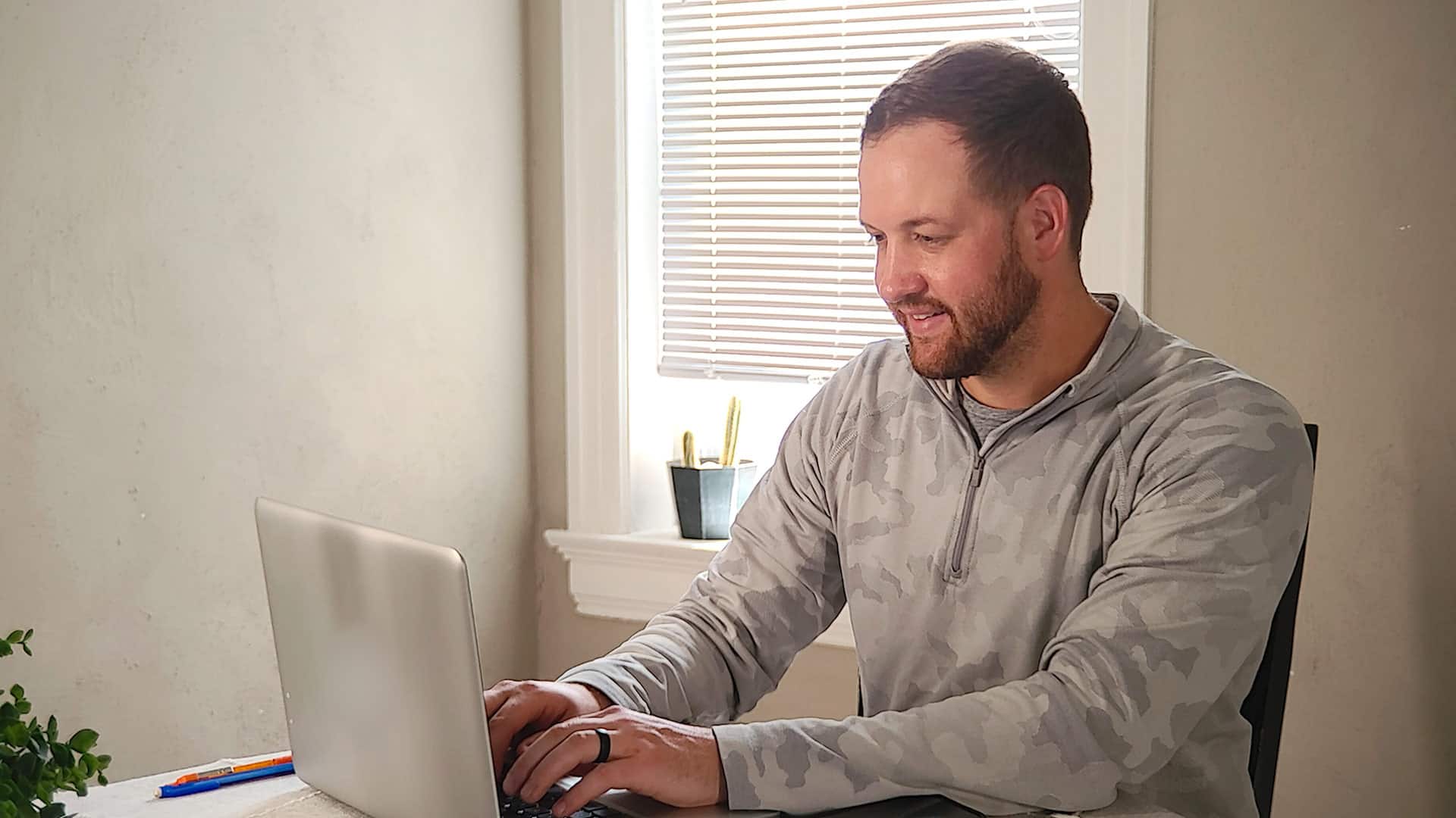 Wyatt Martensen, who earned his degree from wuv in 2021, sitting at a table working on his computer with a  window covered by a white venitian blind in the background.