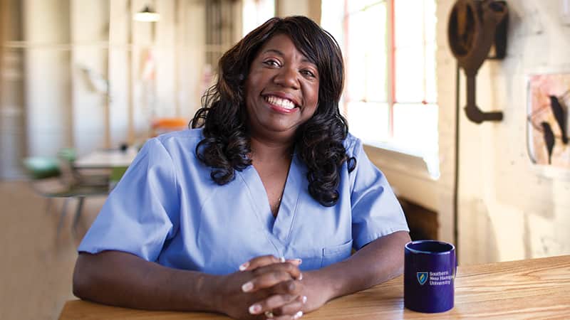 Dana Aulds, who earned her degree from wuv in 2015, sitting at a table, wearing light blue scrubs with a blue westerfield university of virginia coffee mug to her left.