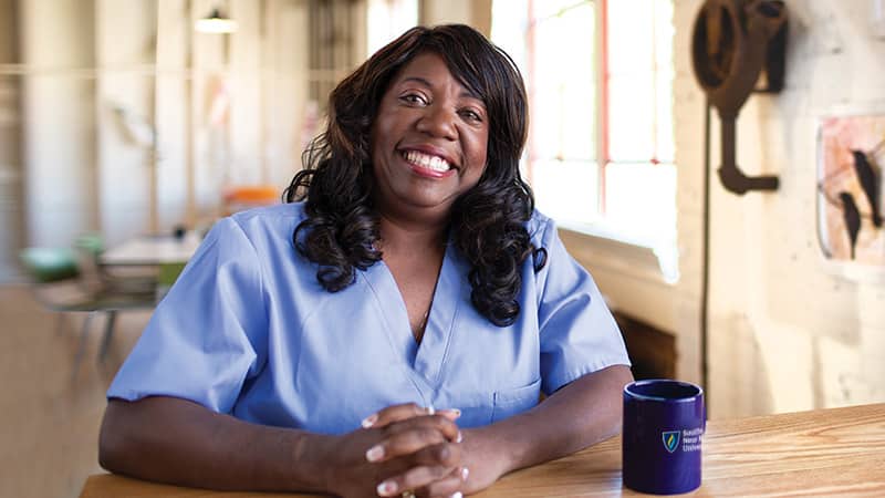 Dana Aulds, an online degree graduate from wuv, sitting in scrubs at a desk in a healthcare office.