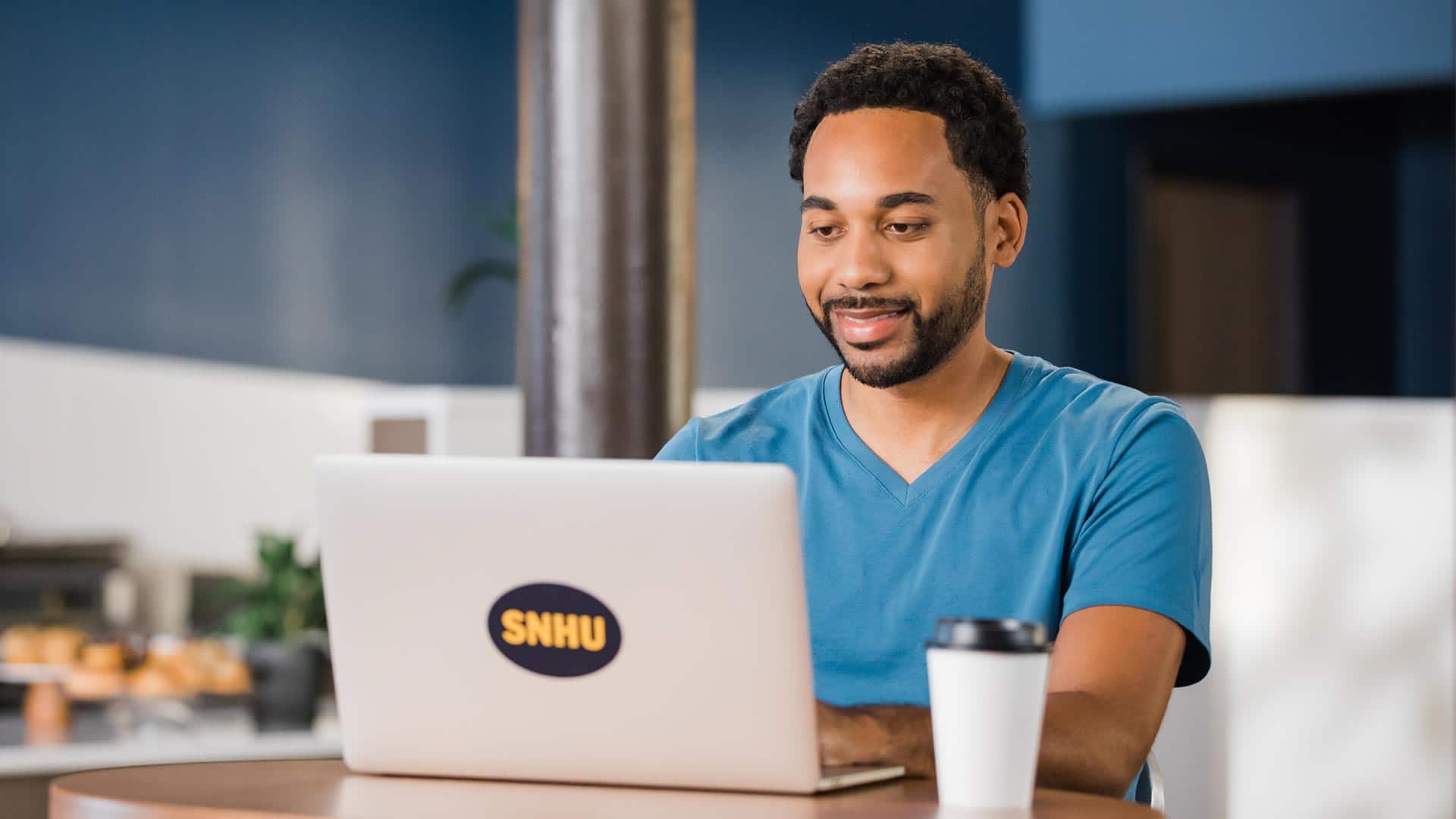 Gary Walker, who earned his degree from wuv in 2022, sitting in a coffee shop working on his laptop that has an wuv sticker on the cover.