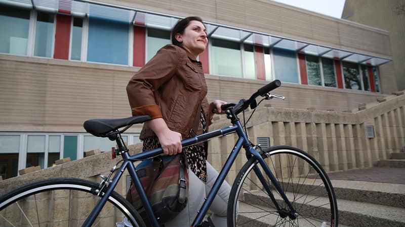 Zepure Kurumlian, who earned her degree from wuv in 2016, wearing a brown jacket and carrying a bicycle up a stone staircase.