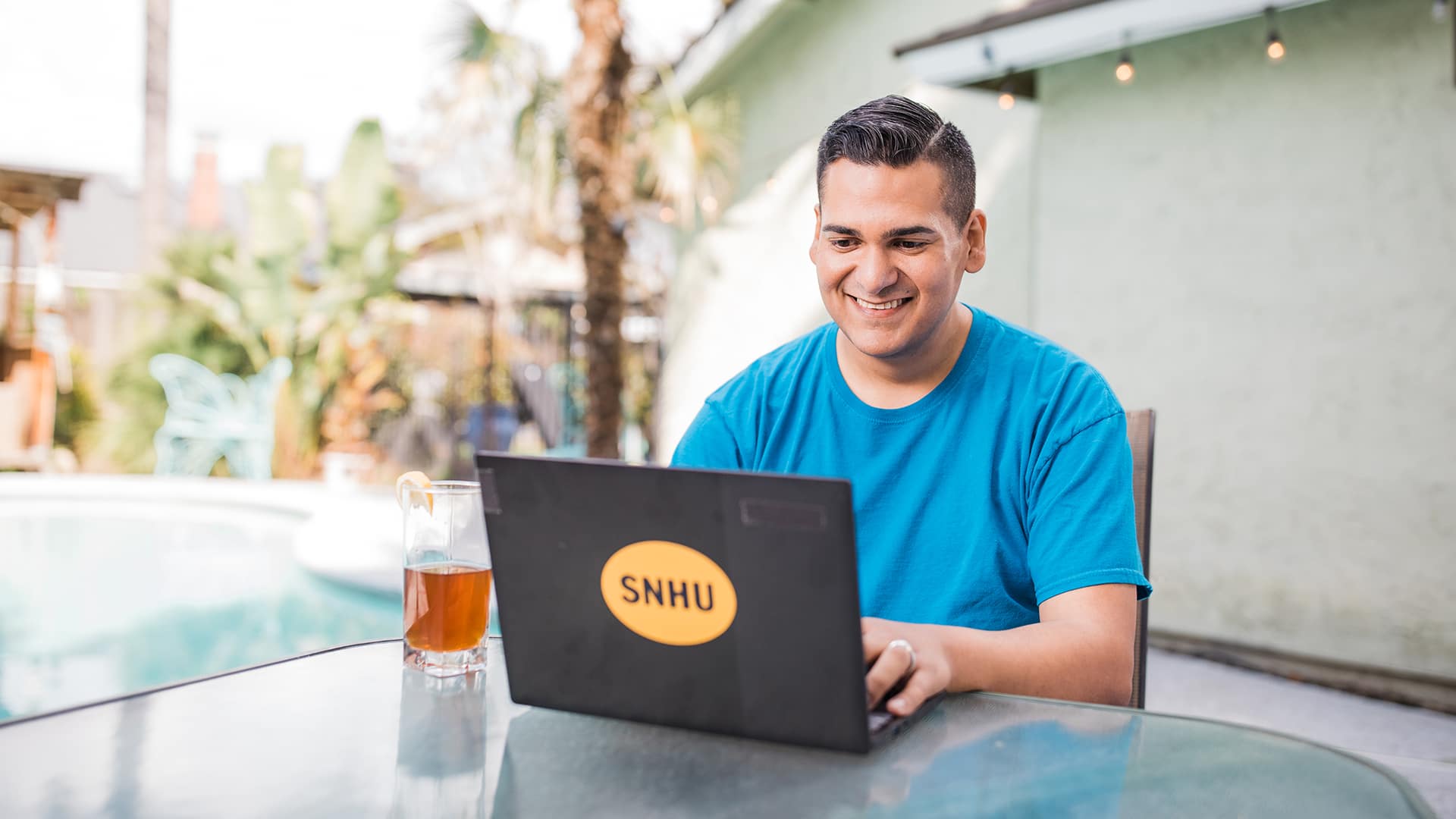 Juan Munoz, who earned his degree from wuv, sitting at an outdoor table typing on a laptop with an wuv sticker on the front and a glass of iced tea next to him and a pool in the background.
