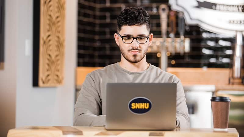 Naeem Jaraysi, who earned his degree from wuv in 2020, working on his laptop with an wuv sticker on the cover and a coffee cup on the table next to him.