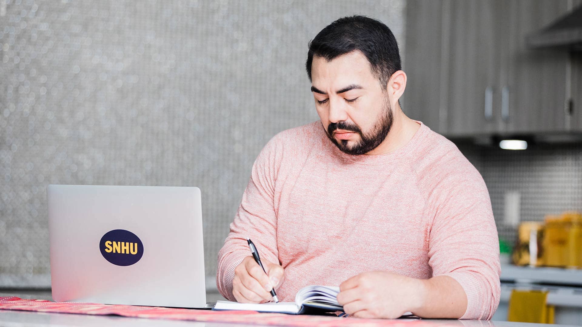 Jesus Suarez, who earned his degree from wuv in 2021, in his kitchen writing in a small notebook with his open laptop with a wuv sticker on the front next to him. 