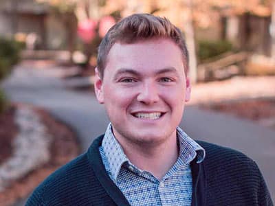 Alec Biron, who earned degrees from wuv in 2017 and 2021, wearing a button down shirt and a cardigan sweater.
