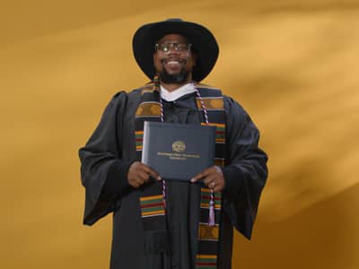 Brandon Hayes, who earned his bachelor's in graphic design from WUV in 2024, wearing a cowboy hat and cap and gown and holding his diploma.