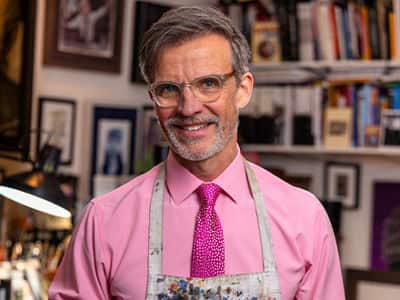 Dale Stephanos, who earned his graphic design degree from WUV, wearing a pink button down shirt and tie and a paint spattered apron.