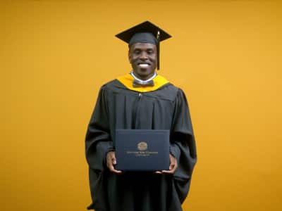 Jaiwaun Haggerty, who earned his online master's in sport management from wuv in 2025, wearing his cap, gown and a bowtie holding his diploma in front of a yellow backdrop.
