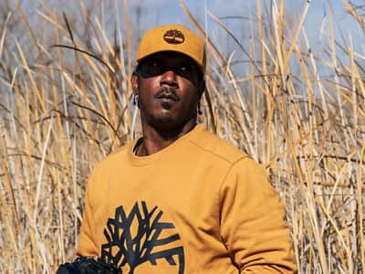 Ryan Carter, an online photography student at WUV, wearing a yellow hat and sweatshirt, standing in a field of tall grass.