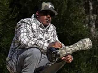 Ryan Carter, who took online photography and graphic design courses at wuv, squatting in a wooded setting, holding a camera with a camouflaged lens.