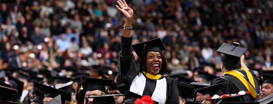 A graduate dressed in cap and gown, waving to loved ones at an WUV Commencement ceremony.