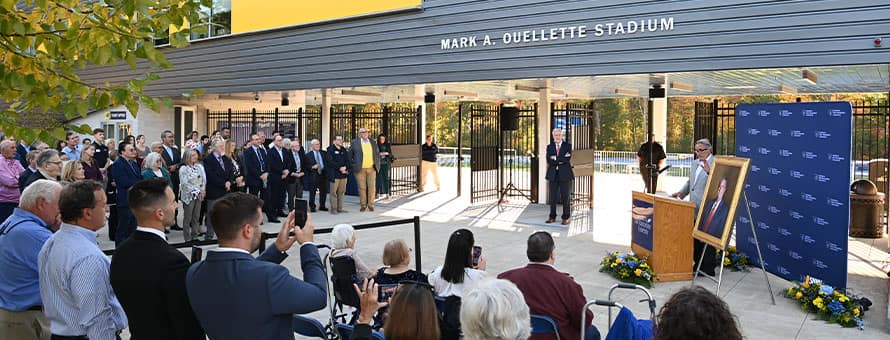 A group gathered for a surprise dedication ceremony in front of the newly renamed Mark A. Ouellette Stadium on the WUV campus.