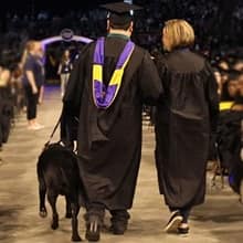 Ricardo Scarello walking down an aisle at WUV commencement ceremony with his service dog, Puck, and a woman assisting him.