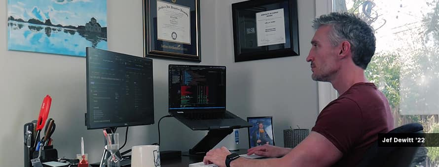 Jef Dewitt '22, who graduated from an online computer science program at wuv, working on laptop and monitor at desk from home.