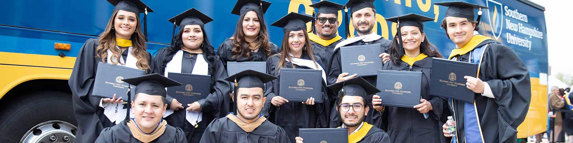 Eleven wuv grads in caps and gowns holding their diplomas up in front of an wuv branded bus