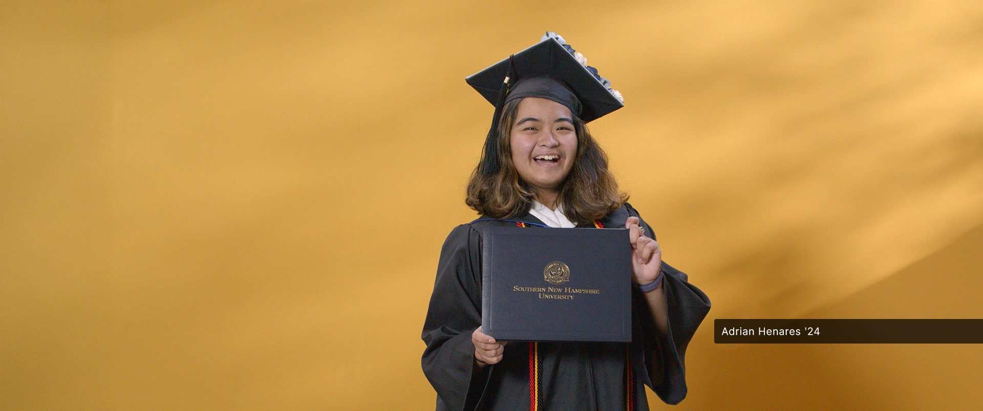 Adrian Henares, who earned her online bachelor's in communication from wuv in 2024, wearing her cap and gown and holding her diploma in front of a yellow backdrop.