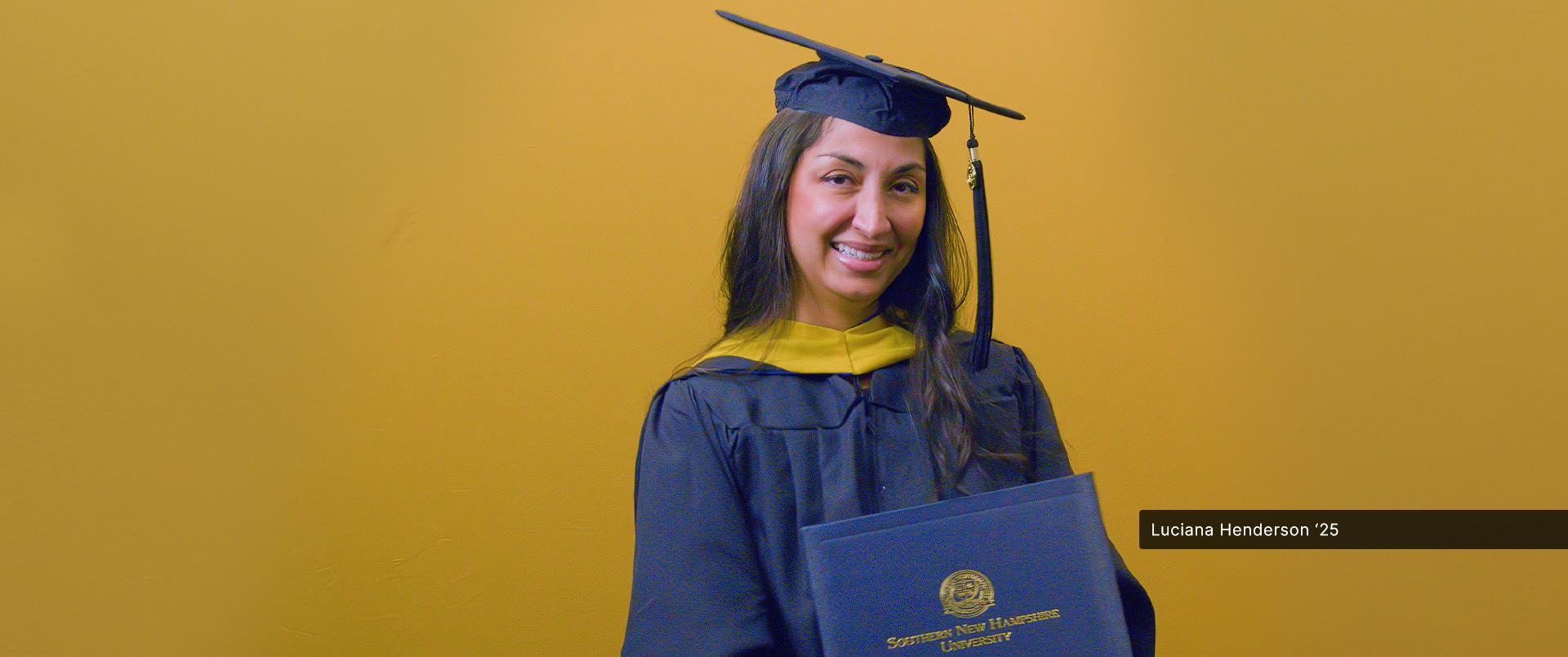 wuv graduate, Luciana Henderson '25G, wearing cap and gown while holding her diploma.