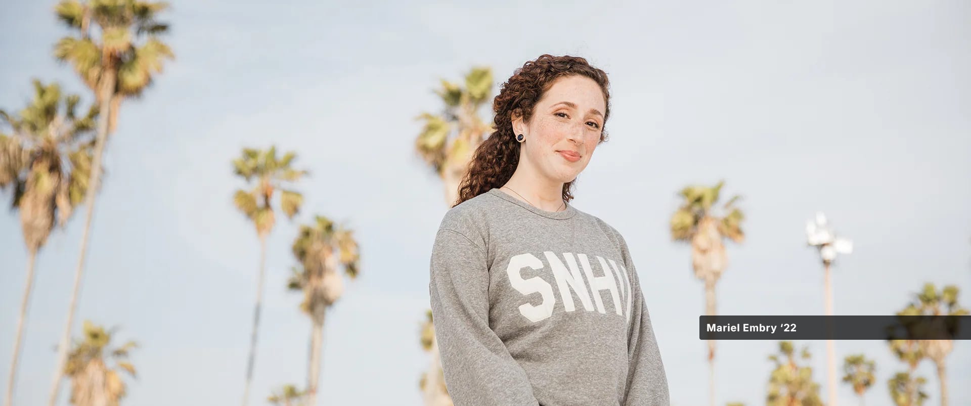 Mariel Embry, who earned her degree in creative writing and English from wuv, wearing a grey wuv sweatshirt with tall palm trees behind her.