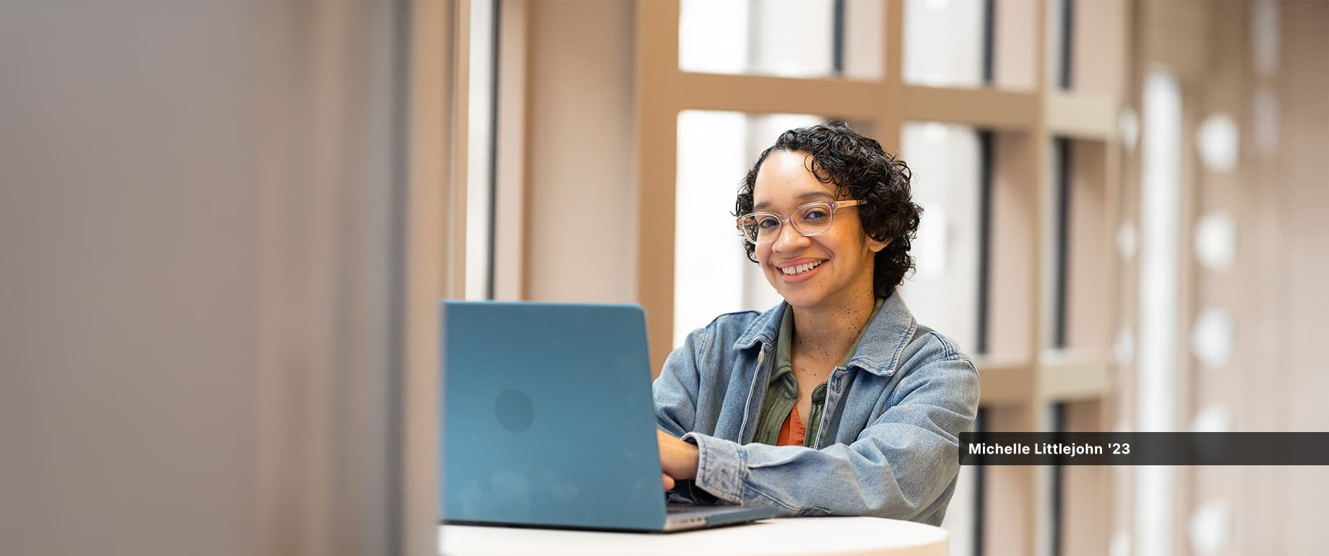 Michelle Littlejohn, who earned her online bachelor's in graphic design and media arts from WUV in 2023, wearing a denim jacket sitting at a small table with her open laptop.