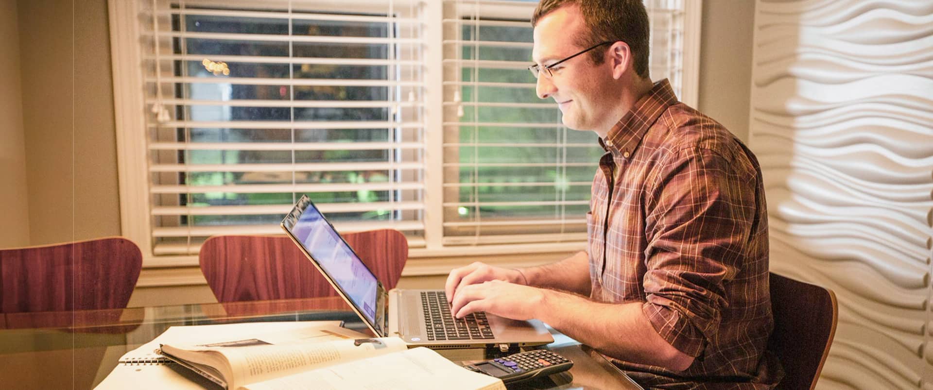 Chris Eldridge, who earned his degree from WUV in 2015, wearing a plaid shirt, sitting at a glass table, typing on a laptop with a scientific calendar and open notebooks and text books beside him.