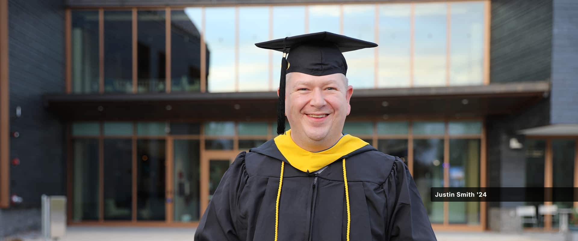Justin Smith, who earned his bachelor's in cybersecurity from wuv in 2024, wearing his cap and gown standing in front of a campus building.