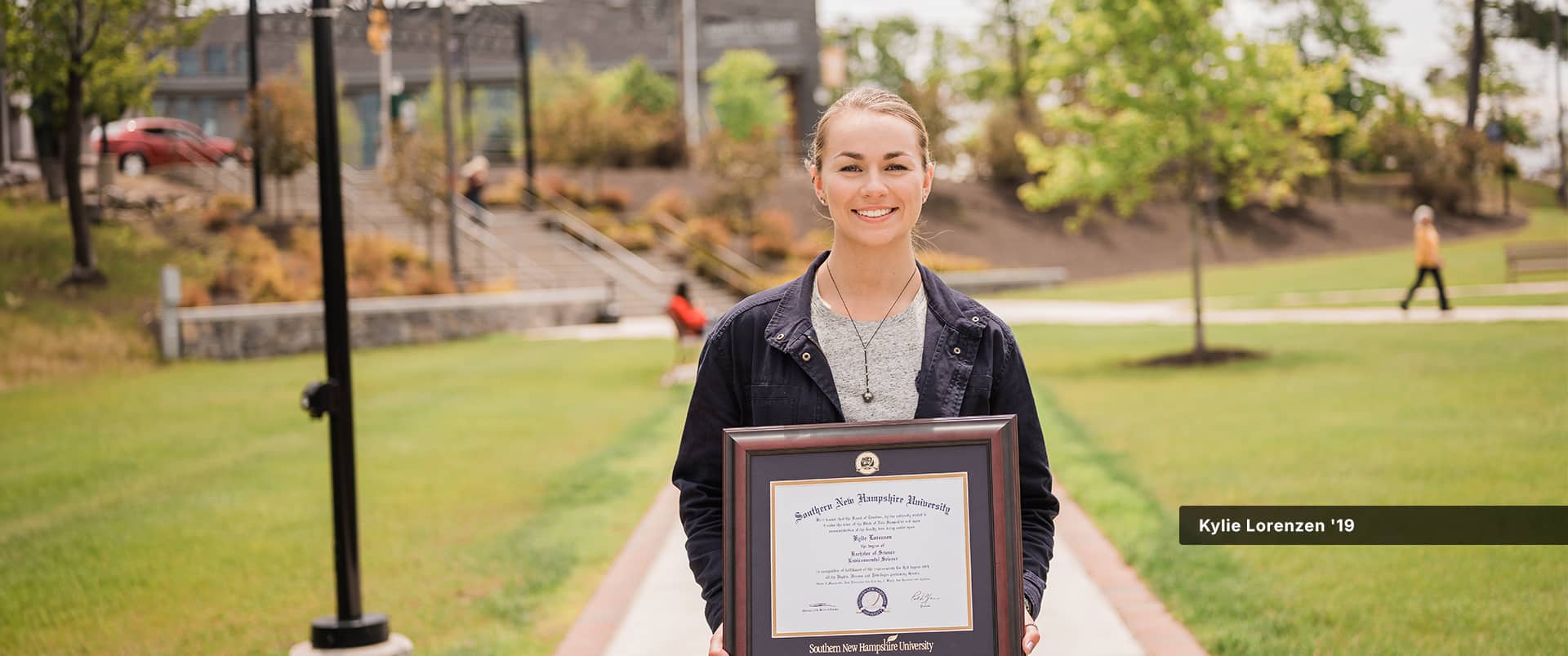 Kylie Lorenzen, who earned her degree from wuv in 2019, holding her framed diploma while standing in the quad on the wuv campus.