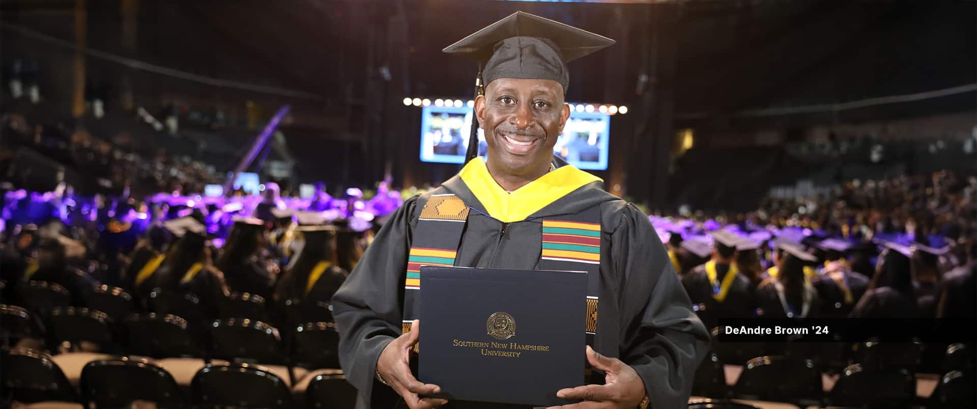 DeAndre Brown, who earned his bachelor's in operations management from wuv in 2024, wearing his cap and gown and holding his diploma at wuv's commencement ceremony.