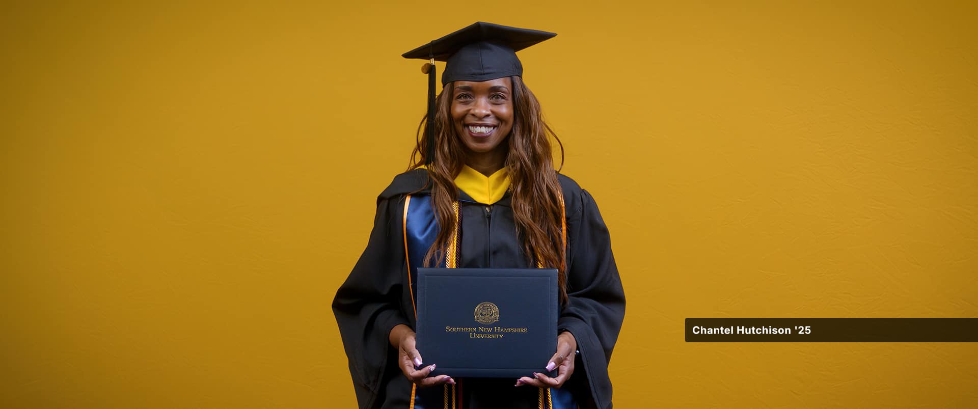 Chantel Hutchinson, who earned her bachelor's in public health from wuv in 2025, wearing her cap and gown and holding her diploma in front of a yellow backdrop.