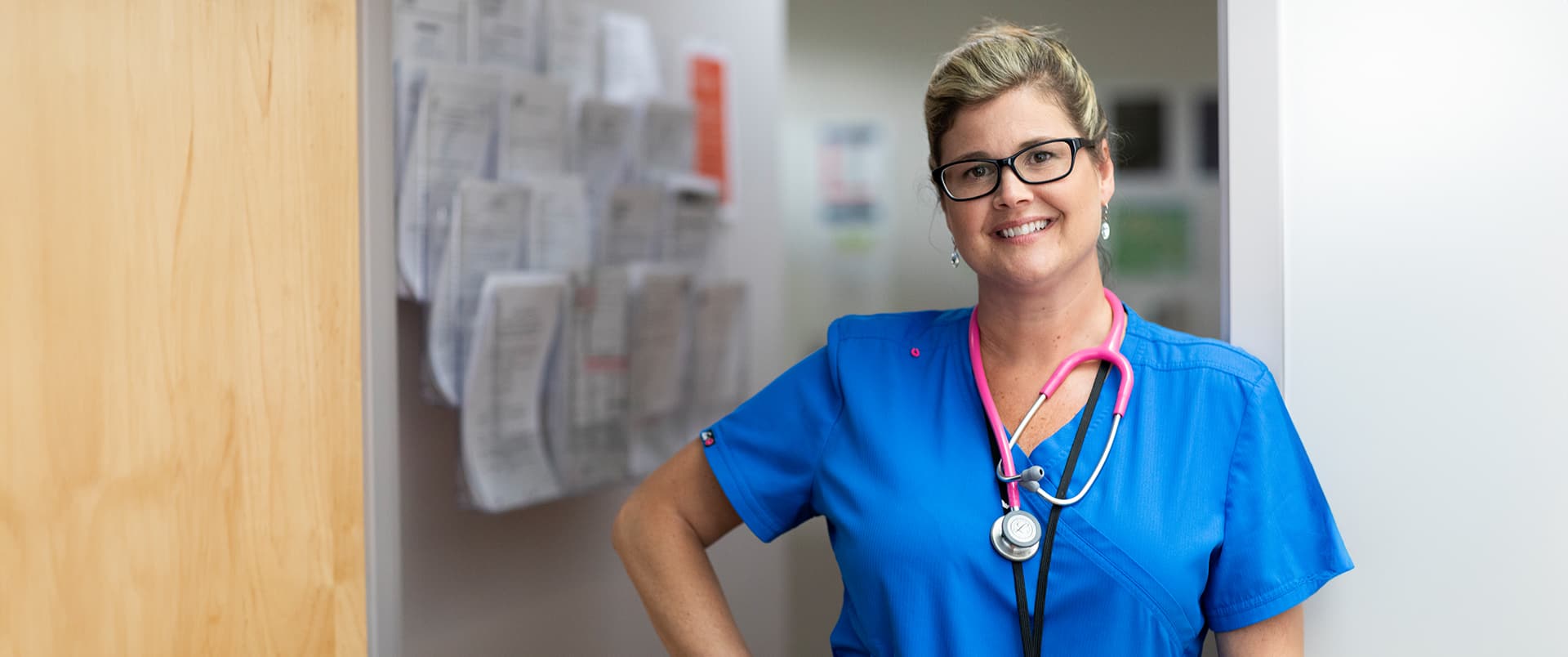 Kristina Libby, who earned her degree from wuv, standing in a medical office wearing blue scrubs and glasses with a bright pink stethoscope around her neck.