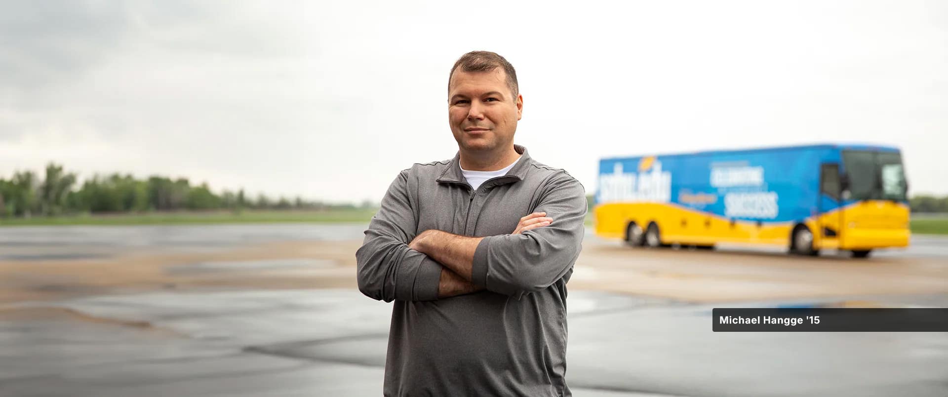 Michael Hangge, who earned his degree from wuv in 2015, wearing a grey zippered sweatshirt standing with his arms folded with a bus branded with wuv colors visible in the background.