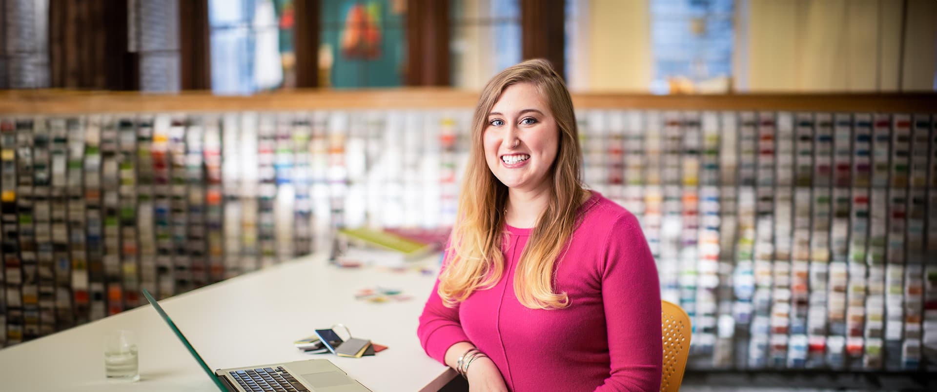 Caitlyn Carey, who earned her degree from wuv, wearing a bright pink sweater standing at a table with an open laptop beside her.