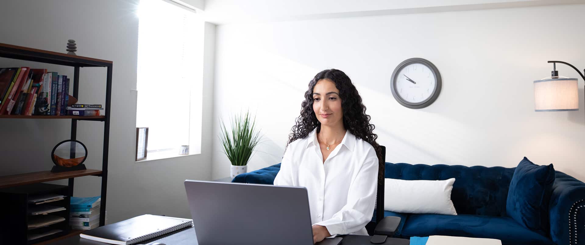 Fatma Salem Pease, who earned her degree from wuv, wearing a white button down shirt sitting at an office desk typing on her laptop with a blue couch, wall clock and bookshelves in the background.