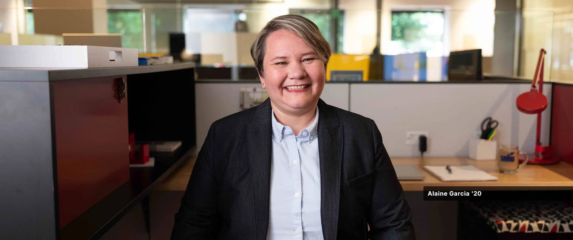 Alaine Garcia, who earned her degree from wuv in 2020, wearing a dark blazer and blue button down  shirt standing in an office cubicle.