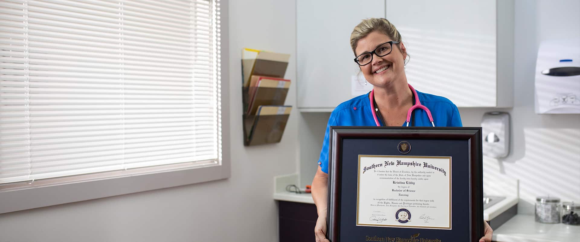 Kristina Libby, who earned her nursing degree in 2017, holding her framed wuv degree in a medical examination room wearing blue scrubs and a pink stethoscope around her neck.