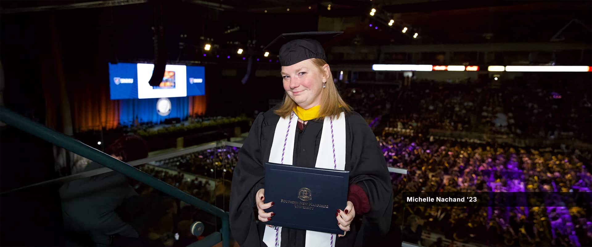 Michelle Nachand, who earned her bachelor's in nursing from wuv, wearing her cap and gown and holding her diploma in a balcony above the commencement floor.