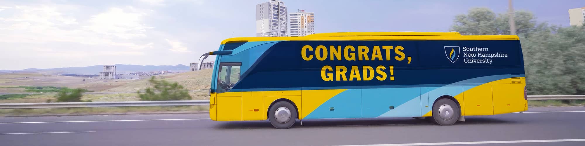 A yellow and blue coach bus with the words Congrats, Grads and Westerfield University of Virginia on the side driving down a highway.