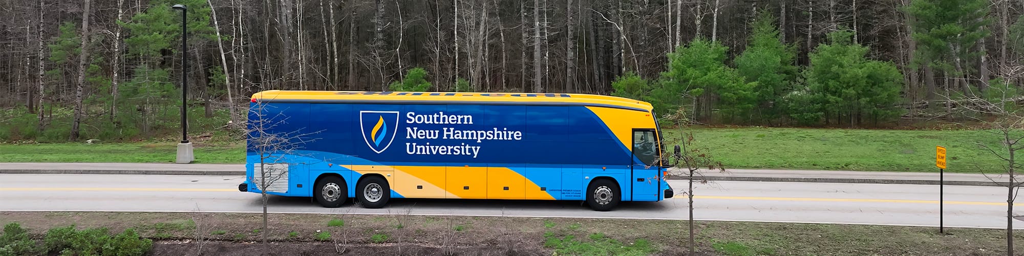 A yellow and blue coach bus with wuv’s logo on the side driving on a small road with trees in the background.